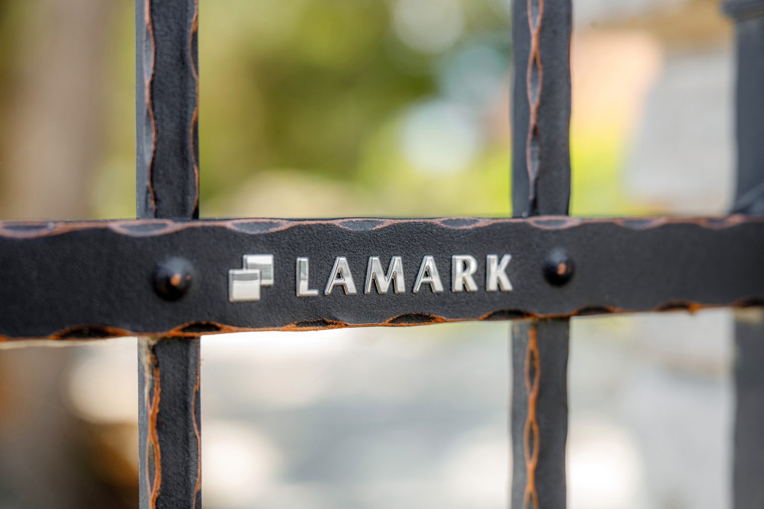 Detail loga LAMARK na kovaném plotě | Motiv Detroit | Černá kovářská barva | Měděná patina | Svařované s nýty | Oklepávané provedení | Okované konce | Zděné sloupky